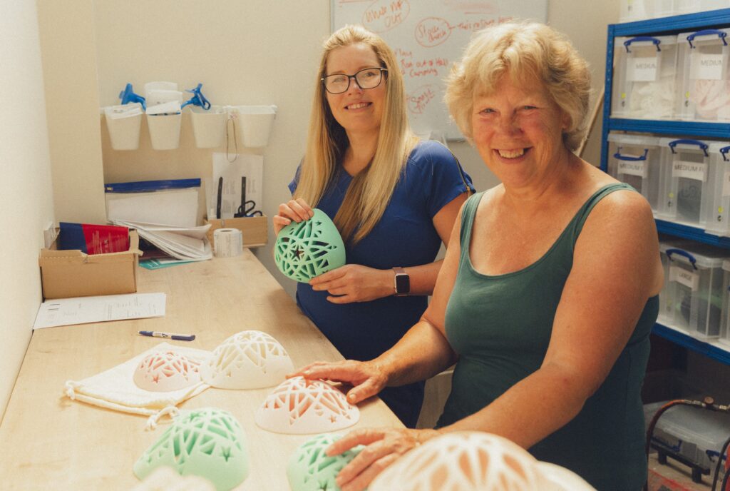 Sam Jackman and her mum standing in a workshop, with breast prostheses on the table 